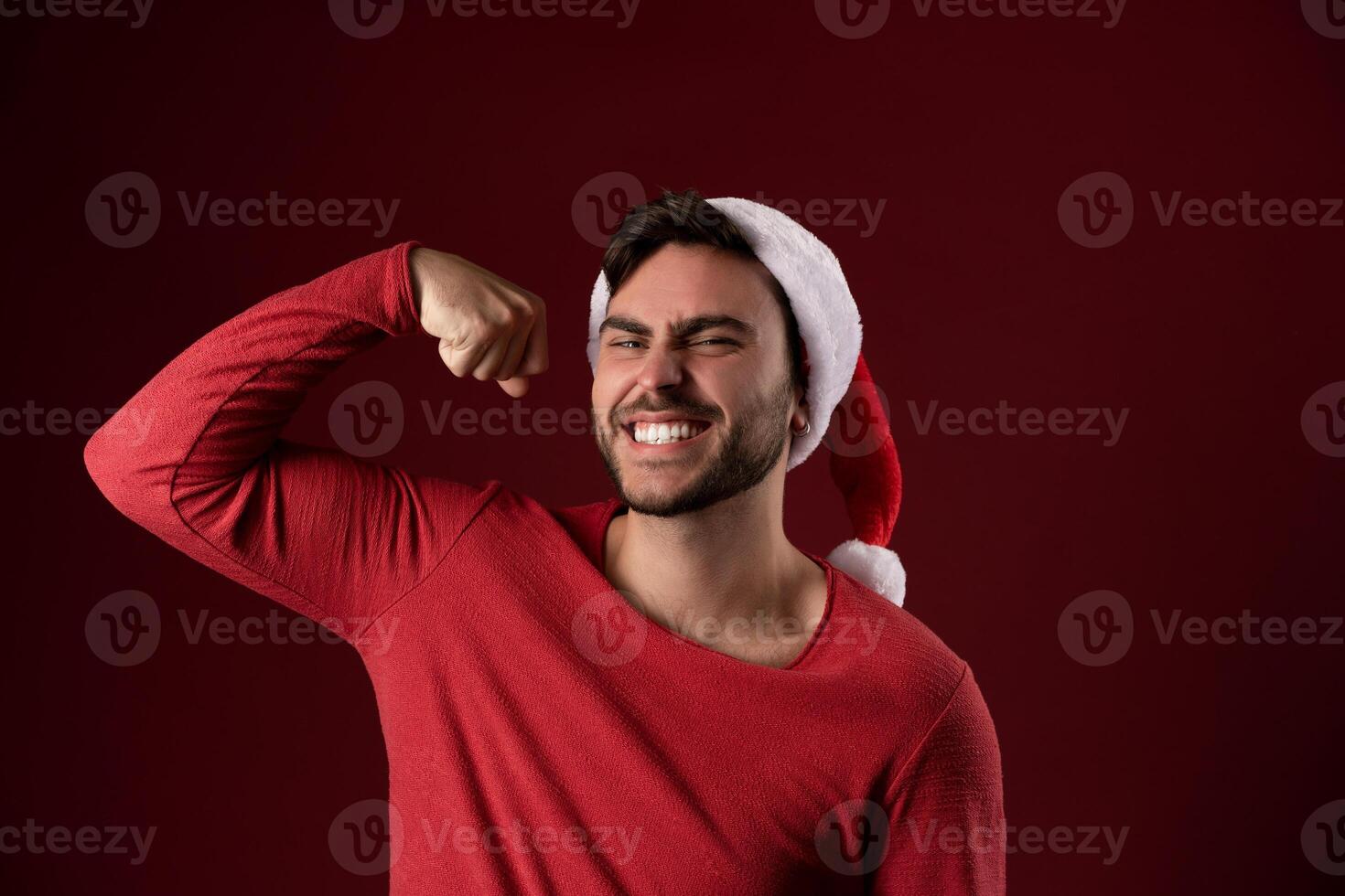 Young handsome caucasian guy in a red sweater and Santa hats stands on red background in studio and showing biceps power photo