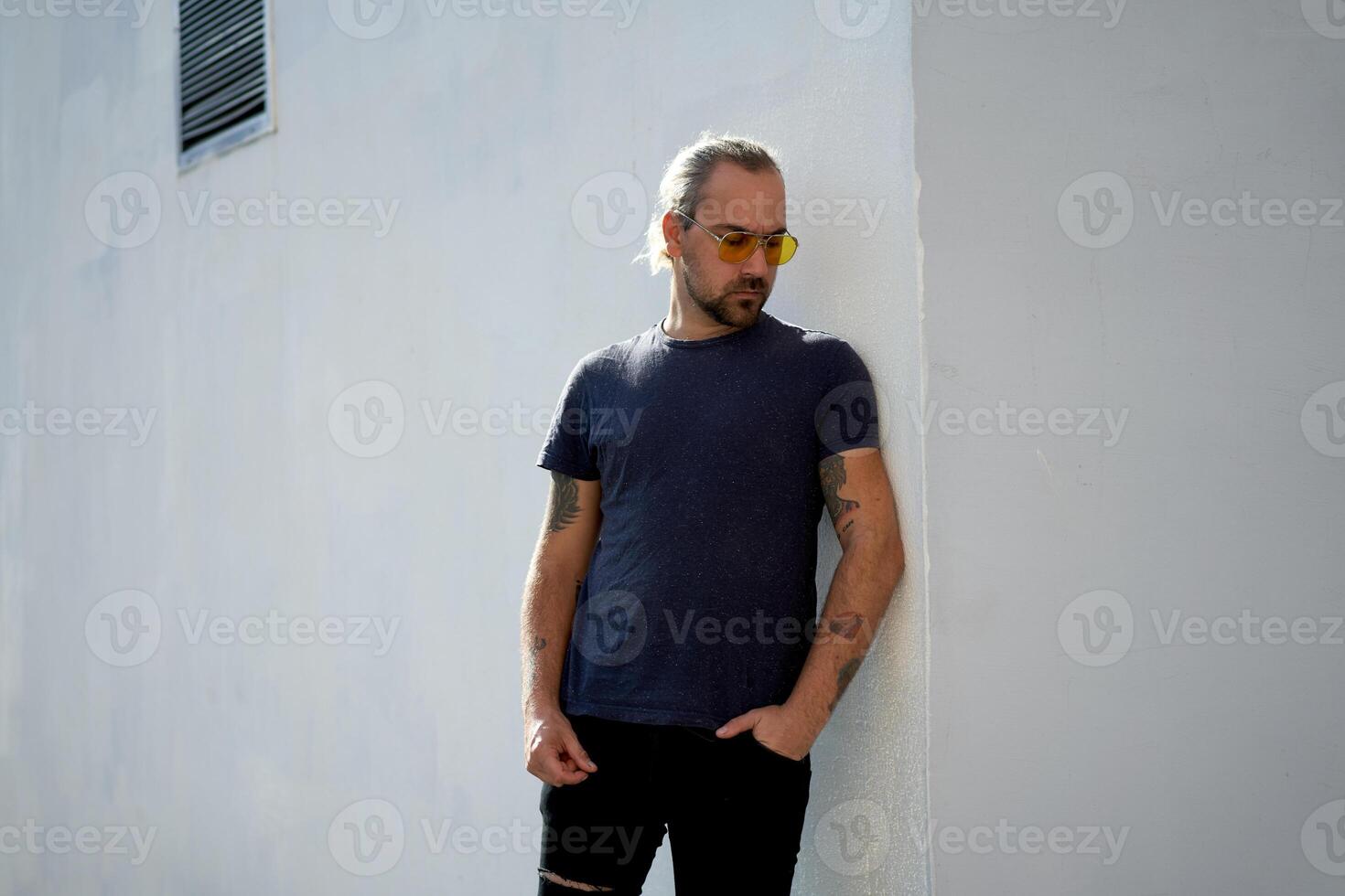 Handsome young unshaven man blue shirt yellow sunglasses stands near white walls summer day. With free copy space for your text photo