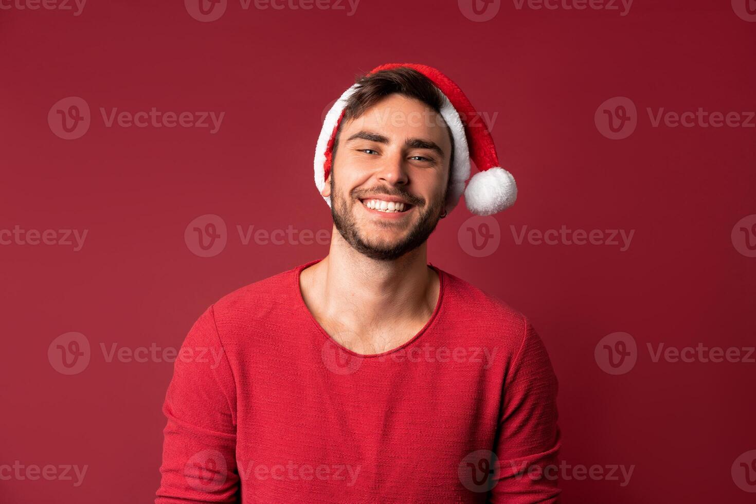 Young handsome caucasian guy in red sweater and Santa hats stands on red background in studio and and teeth smiling photo