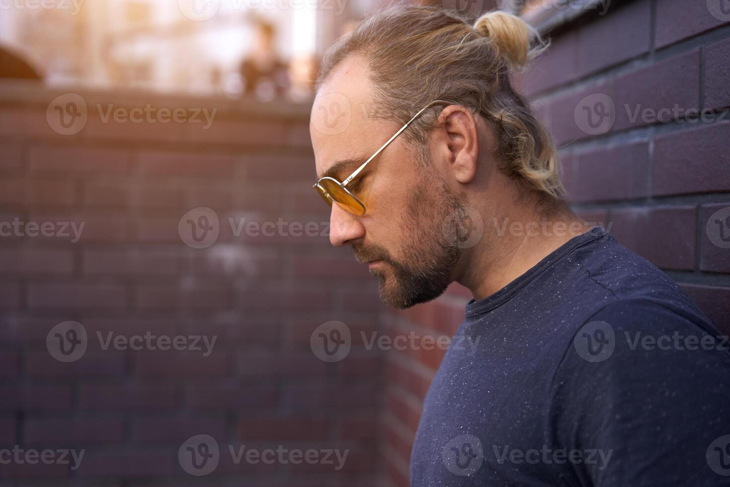 Handsome young unshaven man blue shirt yellow sunglasses stands backyard near brick walls summer day. photo