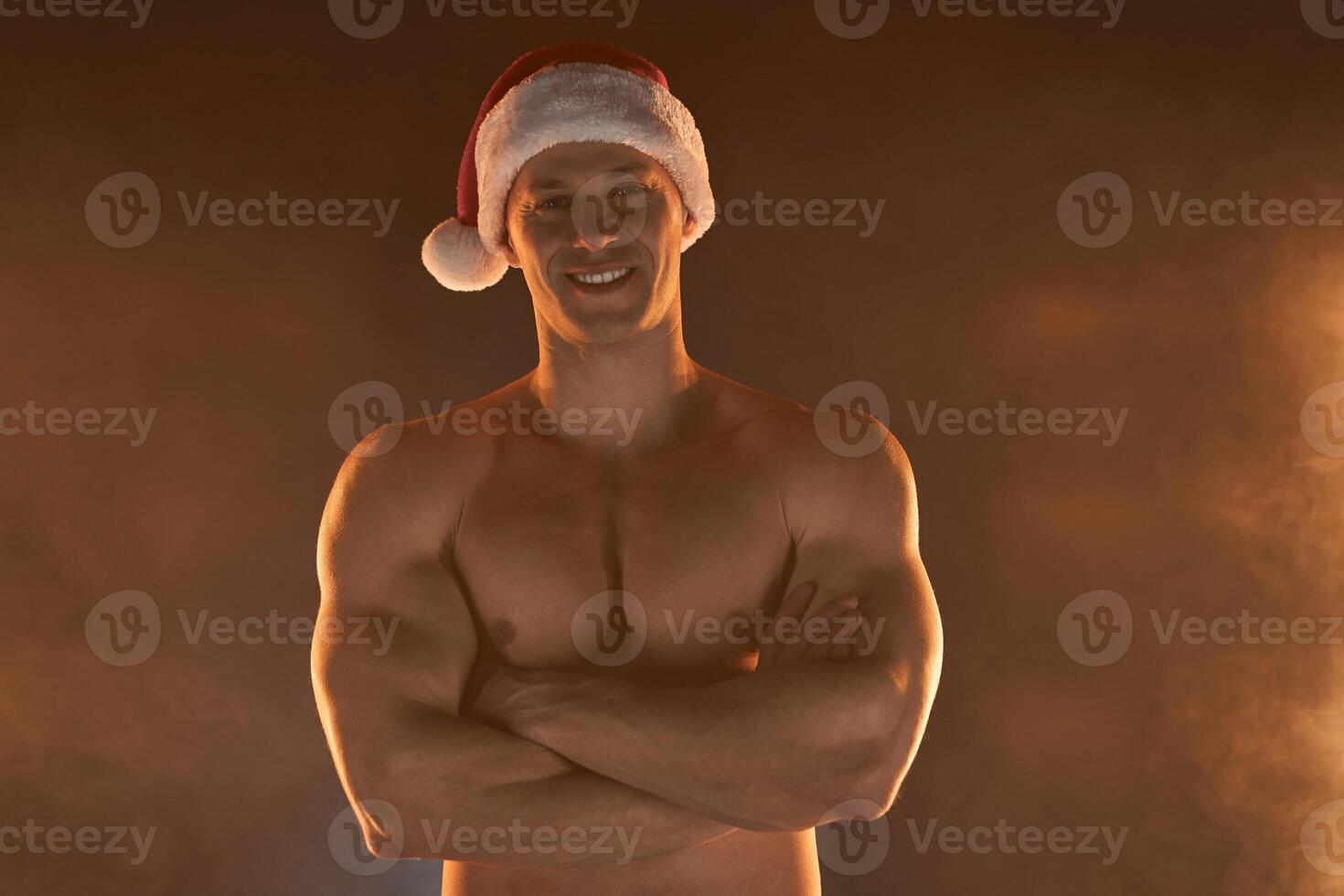 retrato de muscular hombre vistiendo Navidad Papa Noel sombrero, doblada manos en ahumado antecedentes foto
