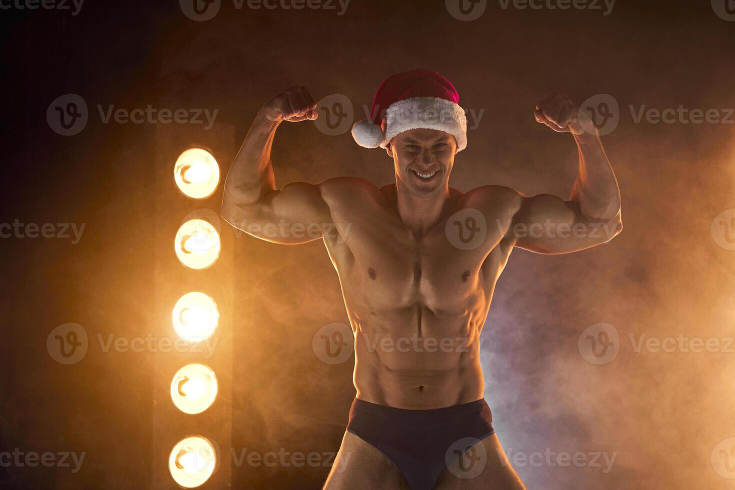 Portrait of muscular man wearing Christmas Santa hat, showing biceps muscle smoky background photo