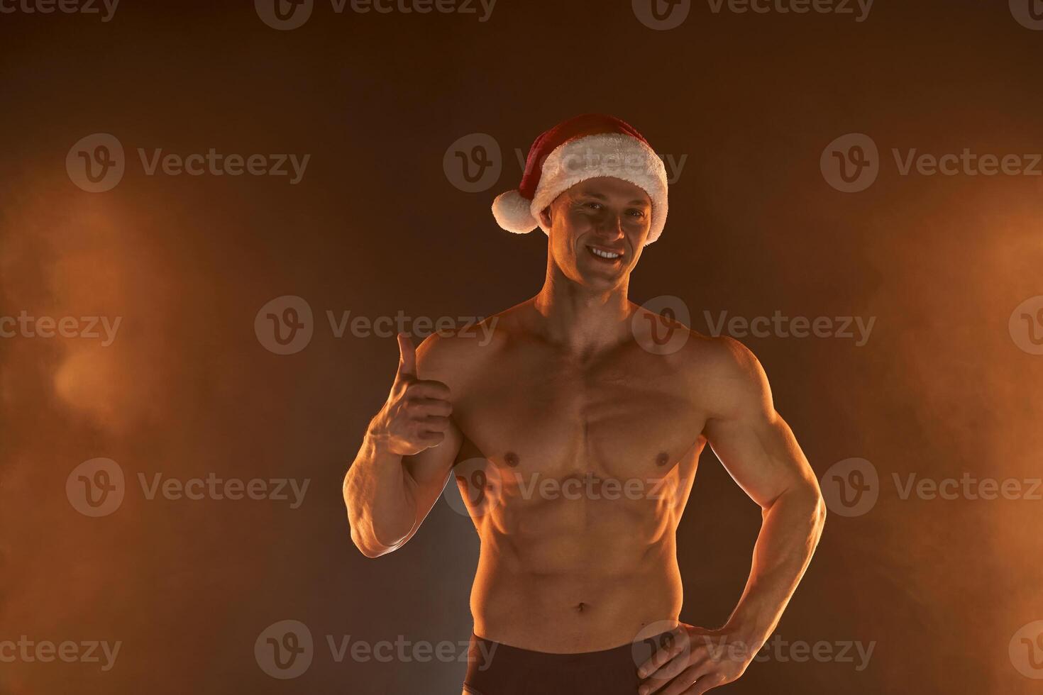 retrato de muscular hombre vistiendo Navidad Papa Noel sombrero, demostración pulgar arriba en ahumado antecedentes foto