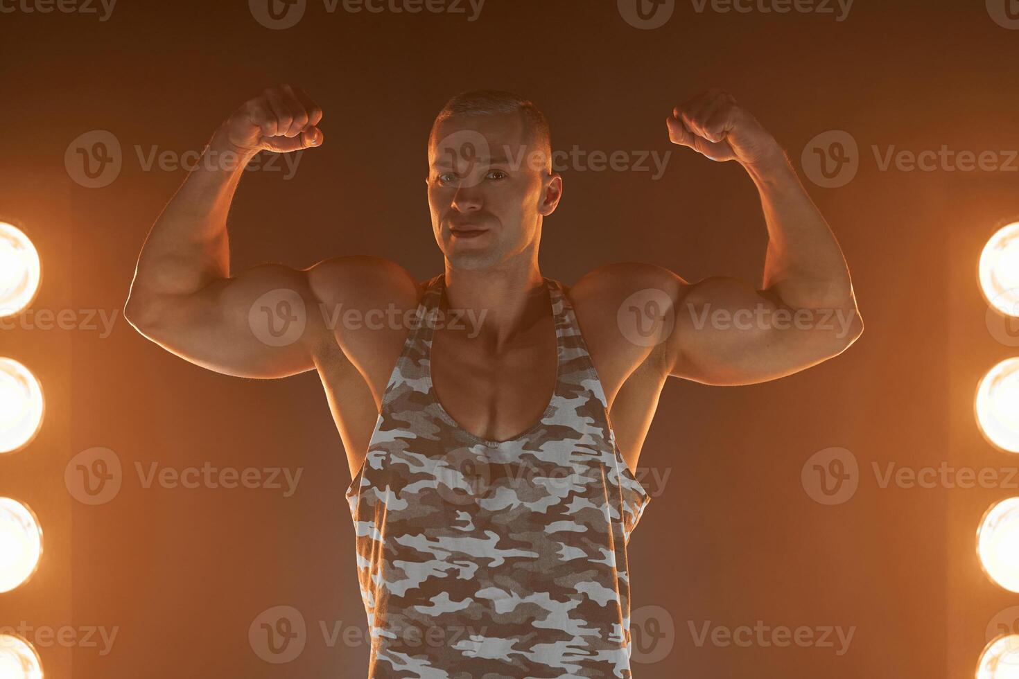 Active lifestyle. Professional bodybuilder showing biceps and smiling at camera, lamps illumination on background photo