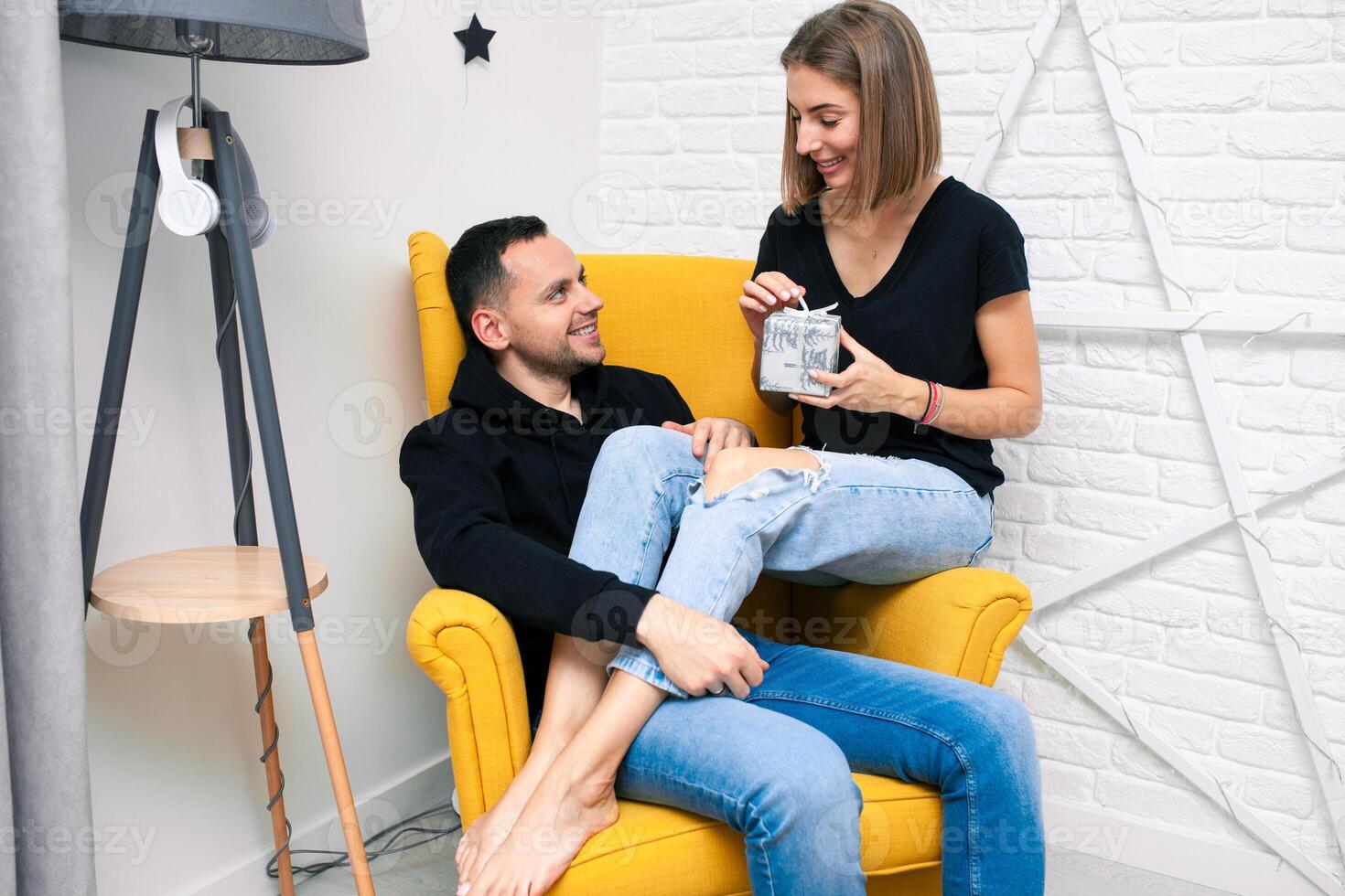 Portrait of couple young in a cozy room. A man sits on a yellow chair. A woman sits on the lap of a man and holds a gift photo