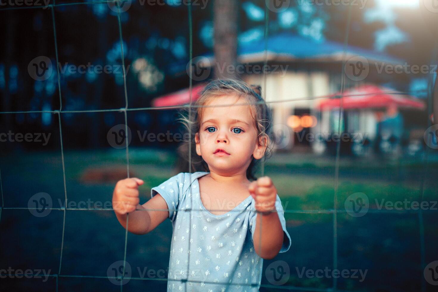 the little girl holds on to the fence with both hands and looks into the camera through the grid photo