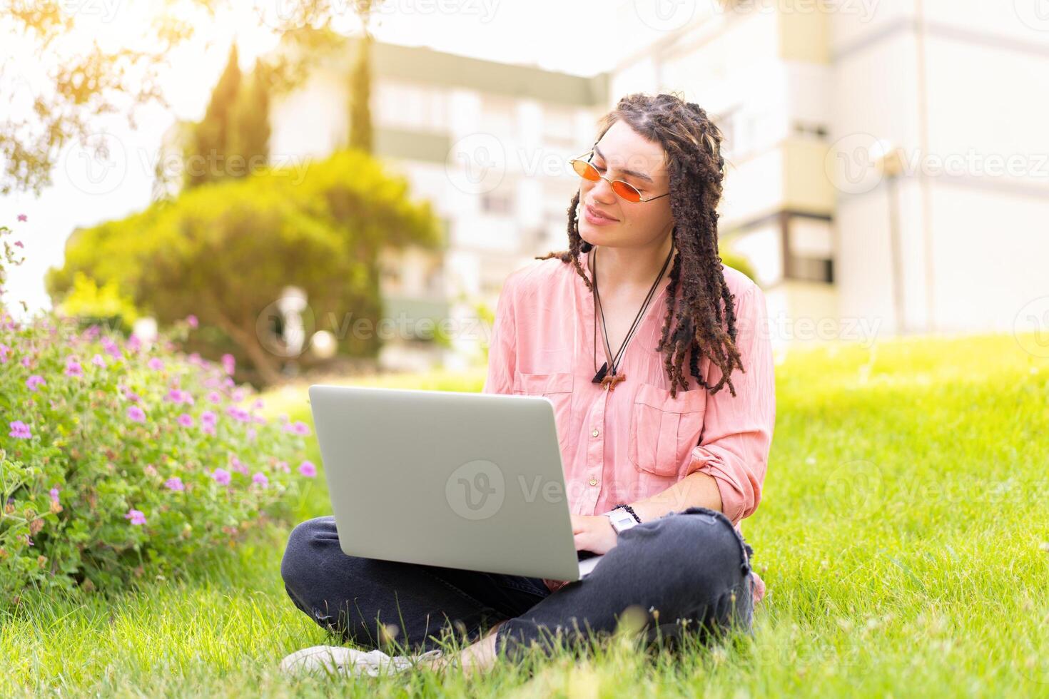 foto de europeo mujer 25 s sentado en verde césped en parque con piernas cruzado durante verano día mientras utilizando ordenador portátil. caucásico hembra hipster con rastas y rosado Gafas de sol en utilizar ordenador portátil