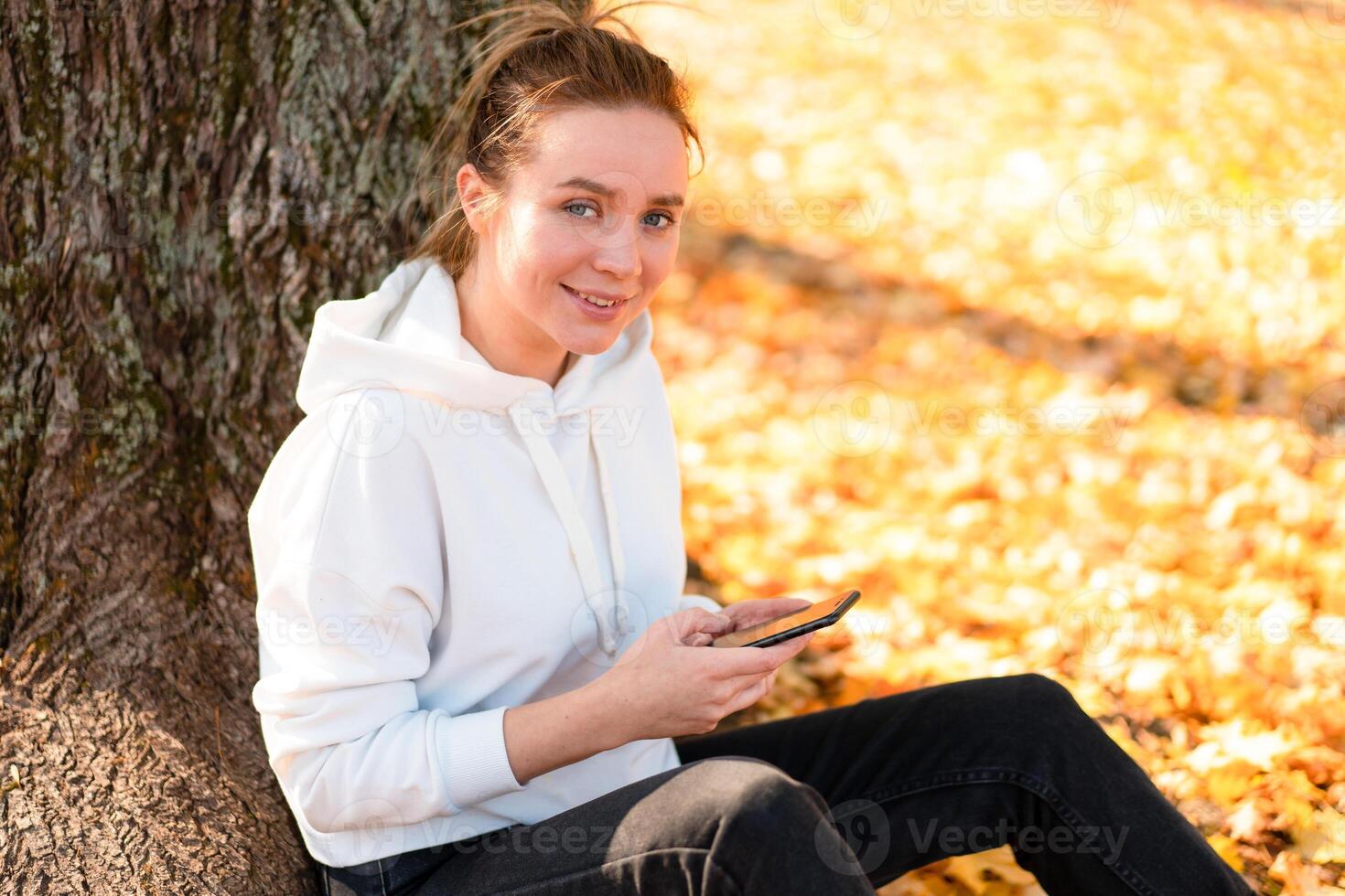 Woman in a white sweater with a hood sits on ground in the park near the tree and holding a cellphone in her hands photo