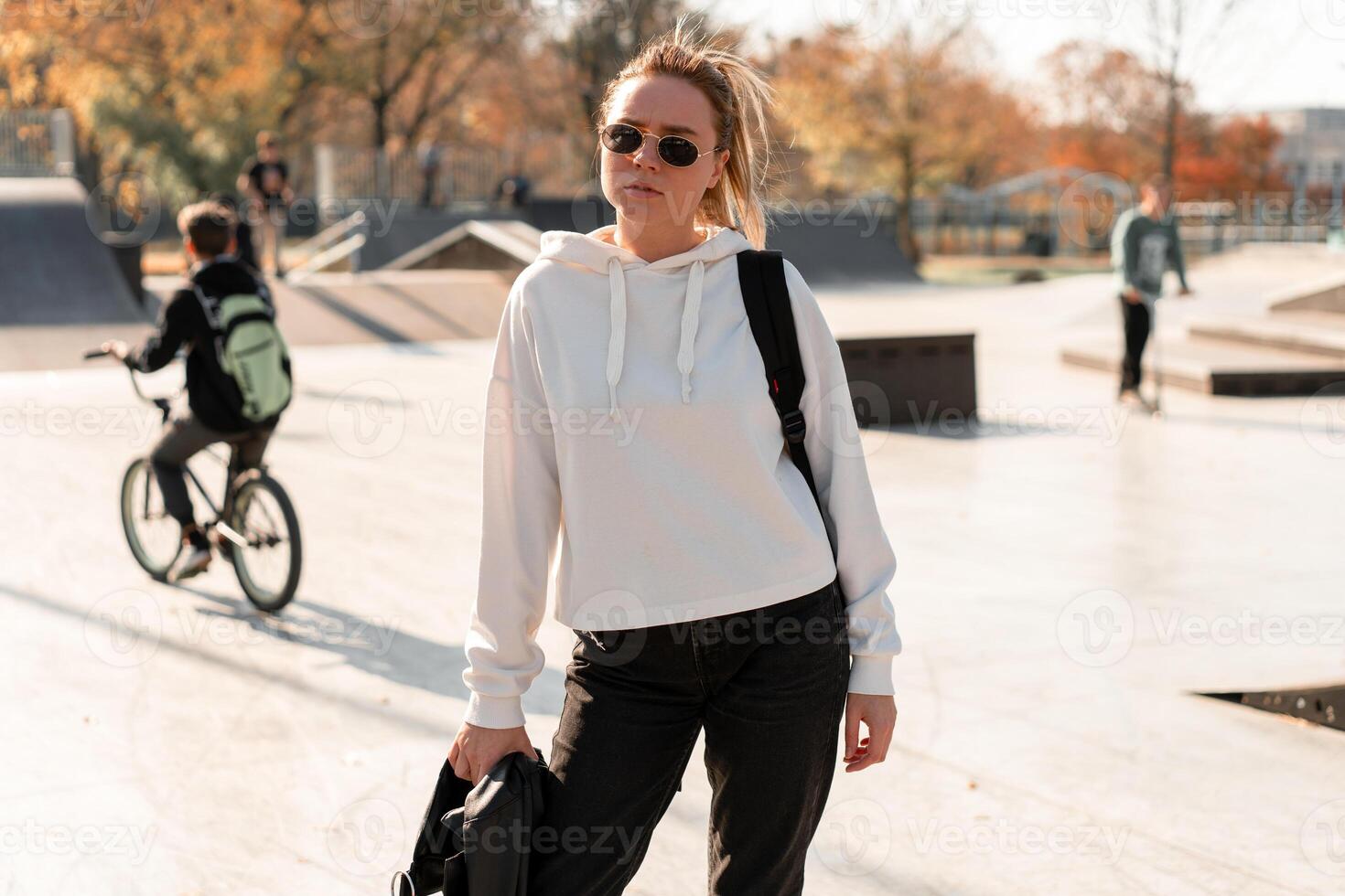 Outdoor portrait of young beautiful woman with a ponytail and sunglasses, with a backpack on his shoulders, dressed in a white sweater, near the sportsground photo