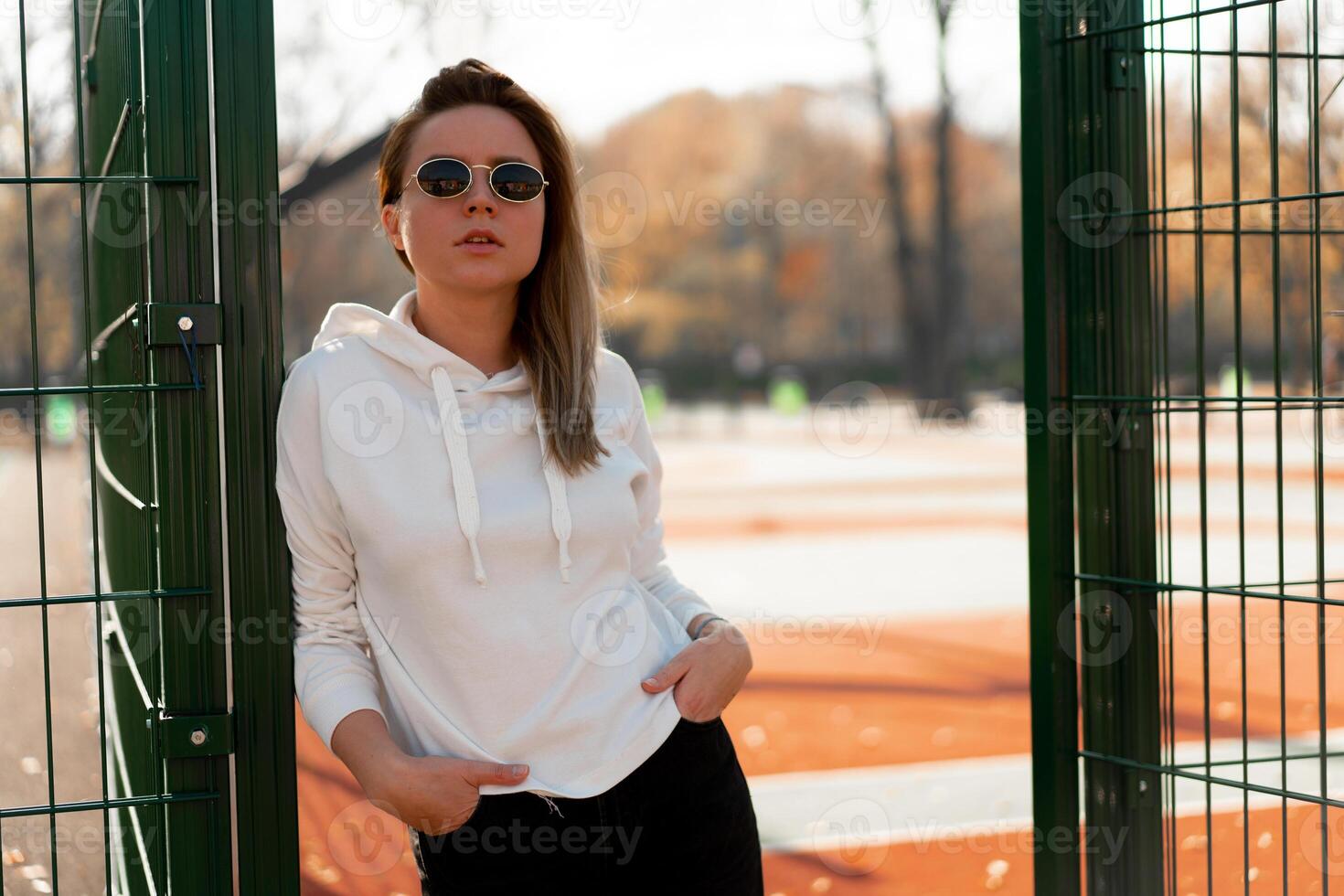 Outdoor close up portrait of young beautiful woman with long hair in sunglasses, dressed in a white sweater, near the sportsground photo