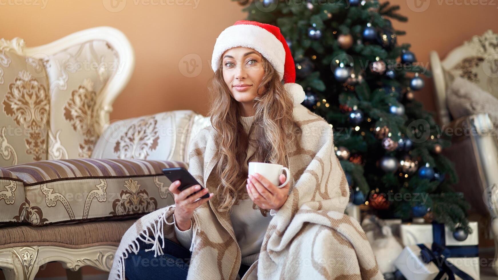 Christmas. Woman dressed white sweater Santa hat on the floor near christmas tree with smartpone and cup of coffee photo