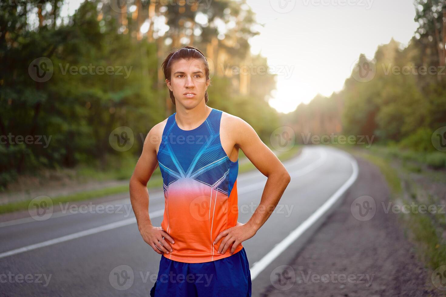Young curly blonde Caucasian man standing on asphalt forest road looking at camera before jogging. photo