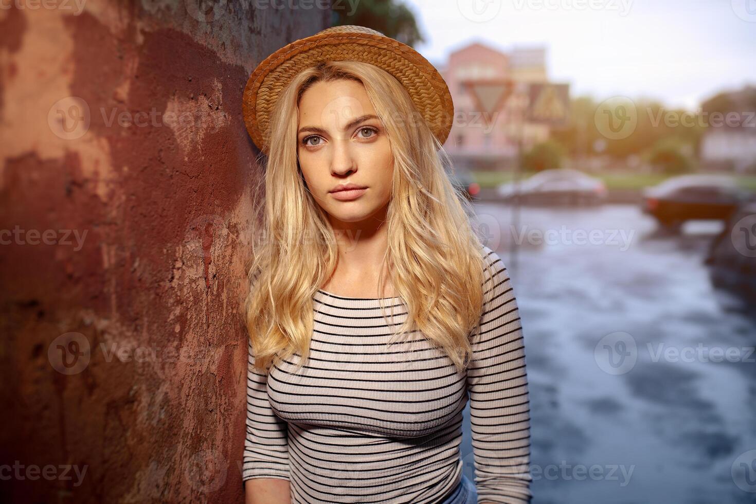 A beautiful young blond female student in a straw hat and a striped blouse is standing alone on the street. The concept of loneliness and sadness photo