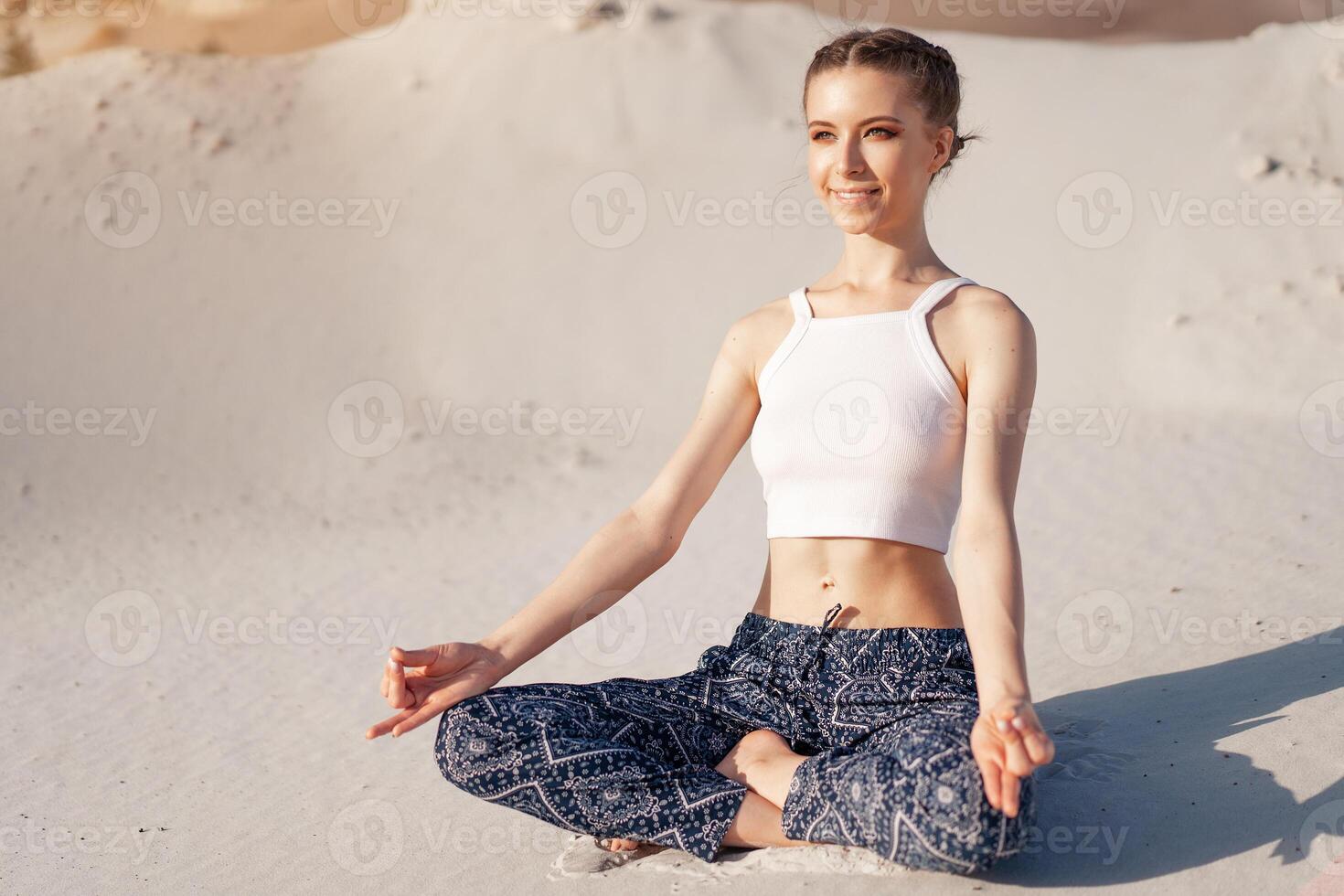 A beautiful young Caucasian girl in a white top and wide pants sits in a lotus position on the beach on the sand. photo