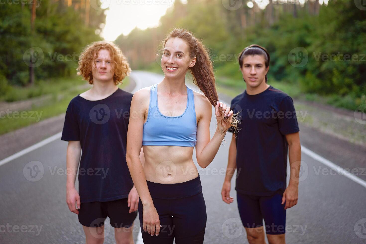 A group of three people athletes one girl and two men standing on asphalt road in pine forest at summer before jogging photo