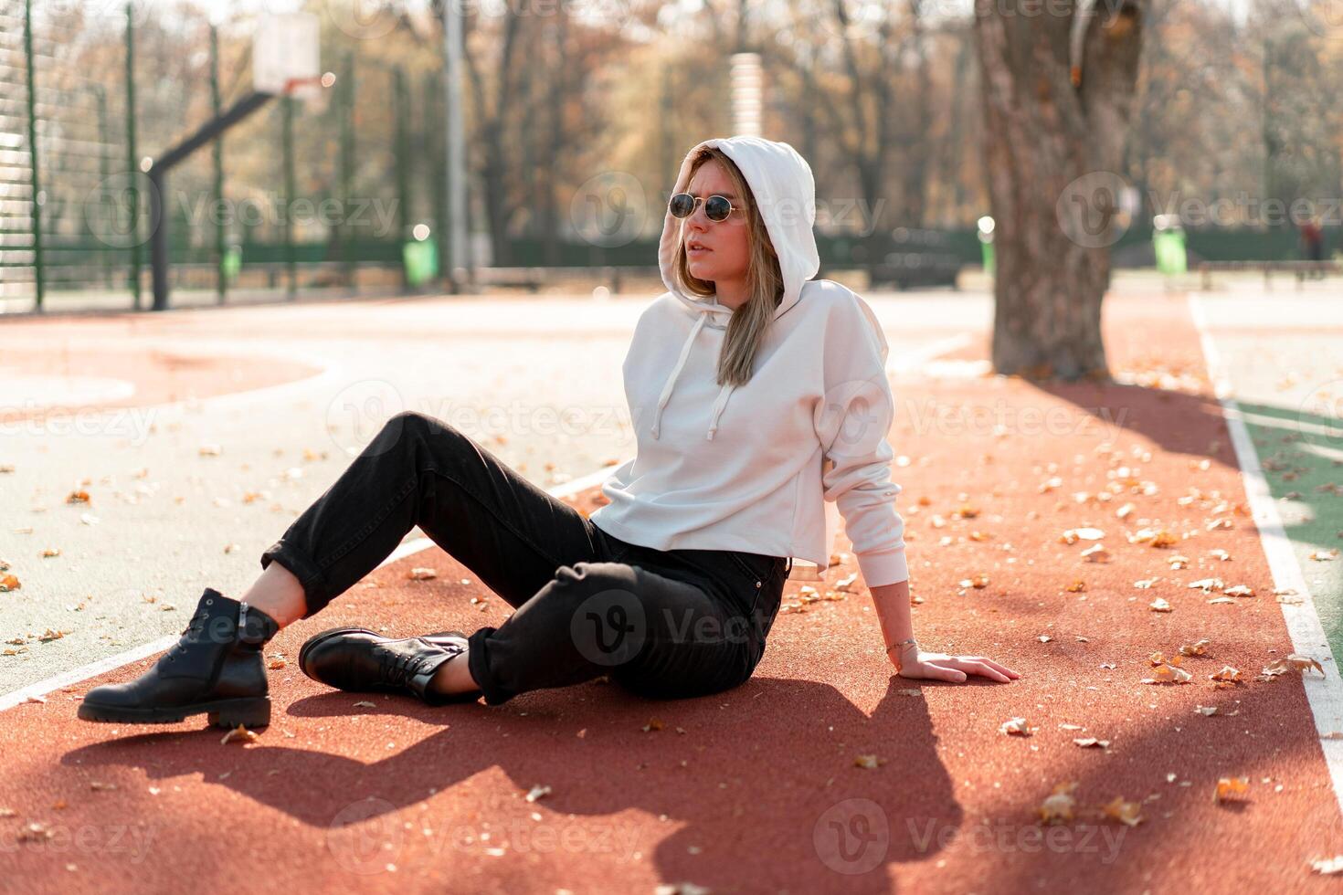 al aire libre retrato de joven hermosa mujer con largo en Gafas de sol y un blanco encapuchado suéter sentado en el campo deportivo pista foto