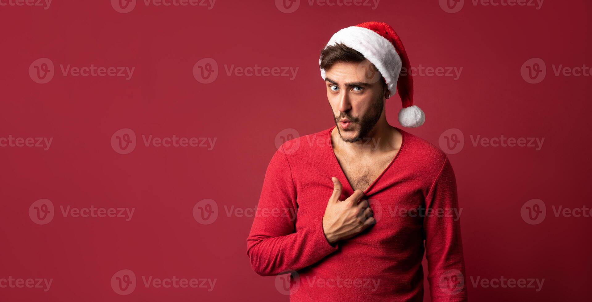 joven hermoso caucásico chico en rojo suéter y Papa Noel sombreros soportes en rojo antecedentes en estudio y muestra cómo caliente él es foto