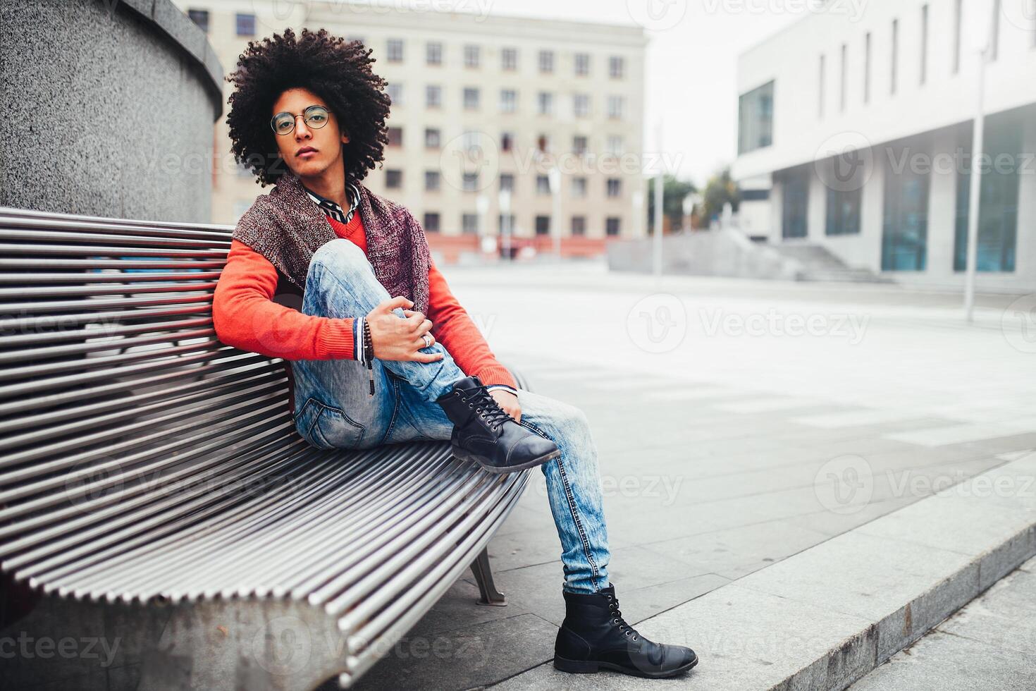 A handsome young Egyptian curly guy sitting on a bench dressed in a bright orange sweater and jeans. The student walks in the break between the lessons. Cute hipster guy photo