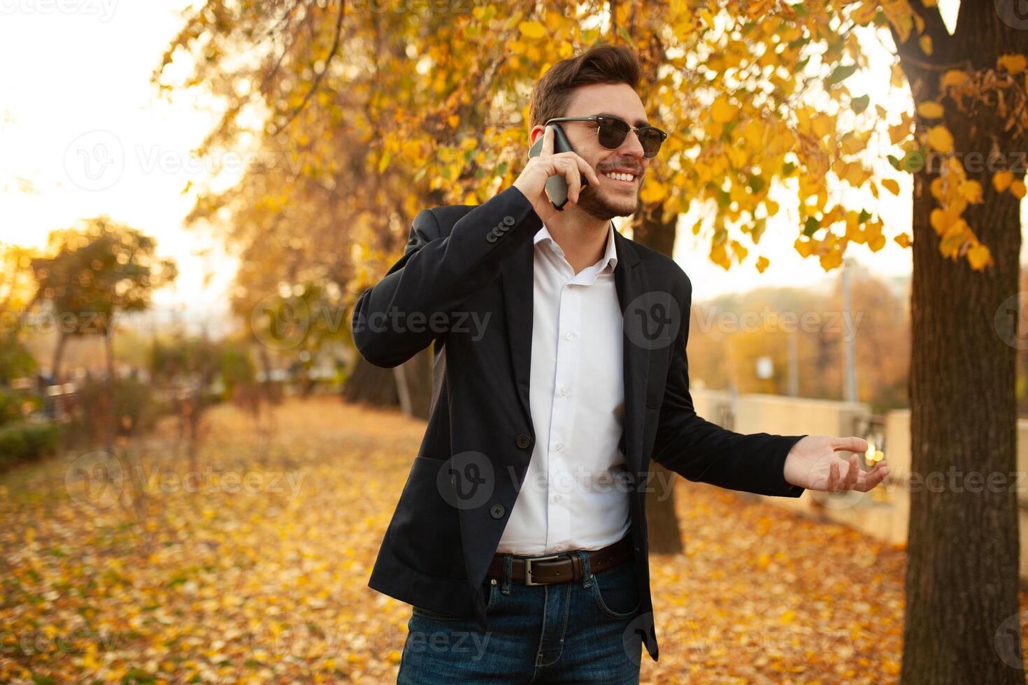 Handsome young stylish male businessman happy and smiling talking on the phone on a walk in the autumn park on the background of yellow leaves photo