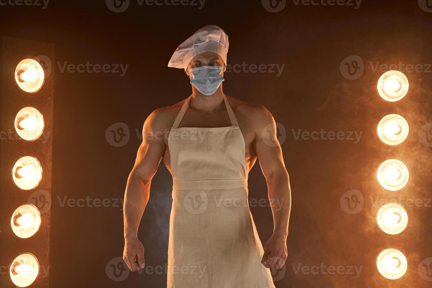 nuevo normal concepto. muscular cocinero vistiendo protector médico máscara delantal y cocinero sombrero, en pie en ahumado antecedentes y lámpara iluminación foto