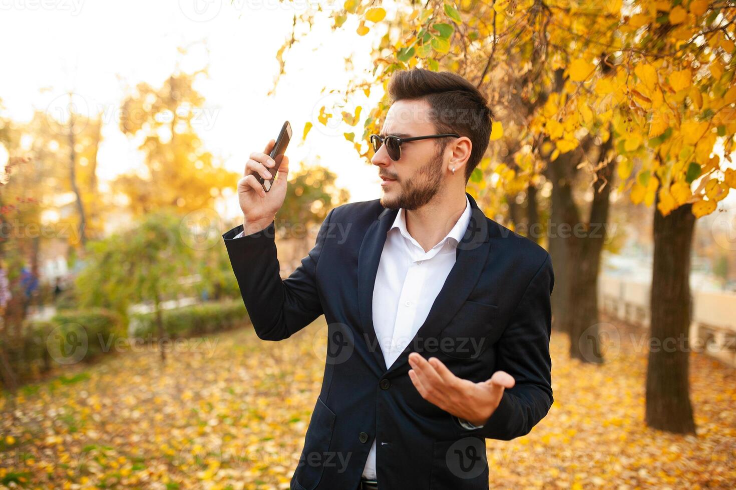 Handsome young stylish male businessman very surprised by unpleasant phone call on a walk in the autumn park on the background of yellow leaves photo