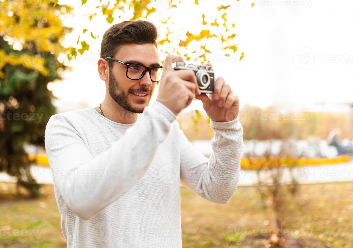 Young handsome hipster guy walks in a beautiful autumn park on the background of yellow leaves in warm sunny weather and takes pictures on a pig film camera. Autumn leisure time. Creative youth photo