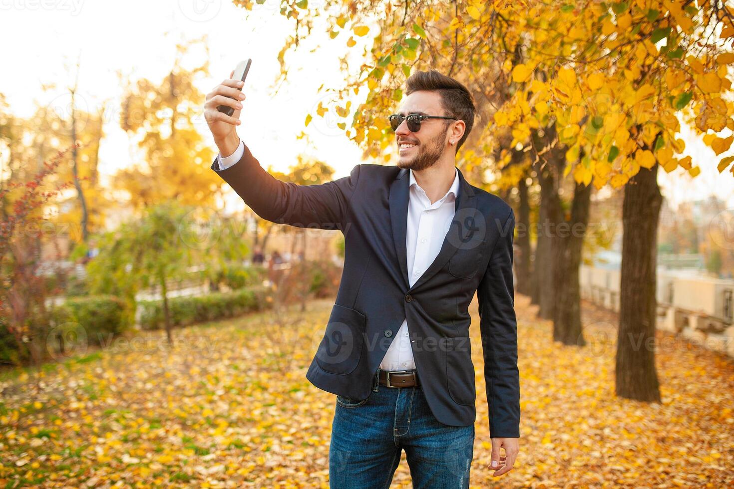 Handsome young fashionable man in a stylish business suit and sunglasses do selfie in his phone walking in the autumn park on the background of bright yellow leaves. photo