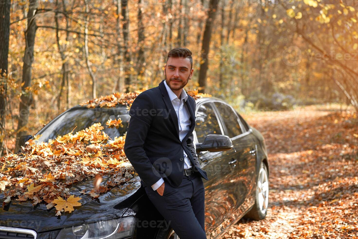 A handsome young Caucasian man wearing business suit standing near black car on nature. photo