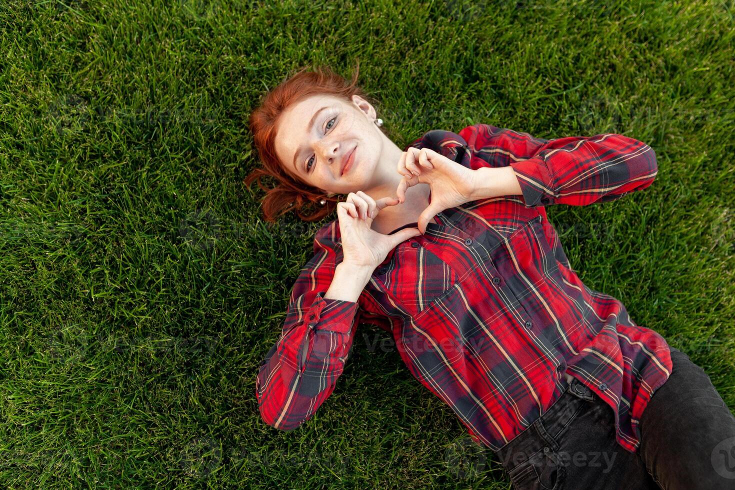 Beautiful red-haired young student with freckles lying on her back on the grass top view. Dressed in a red checked shirt, reveals a heart of fingers Love and positive photo