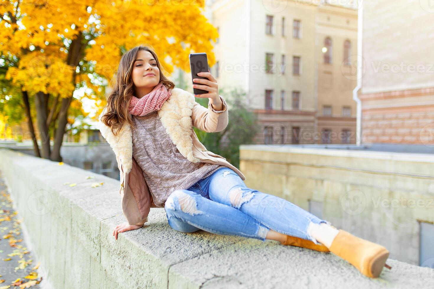 Beautiful caucasian brunette girl sitting warm autumn day with background of trees with yellow foliage and a city. Dressed wool sweater and jacket. photo