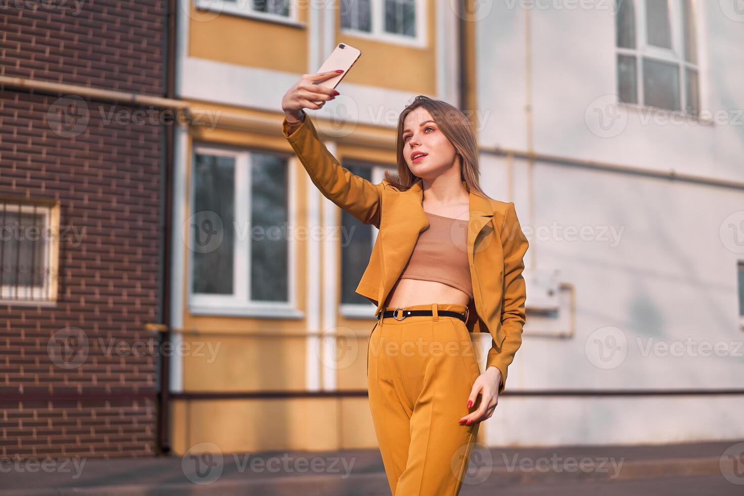 hermosa joven caucásico niña 20 años antiguo con blanco pelo vestido elegante naranja negocio traje hace selfie en el teléfono mientras en pie en el calle. foto