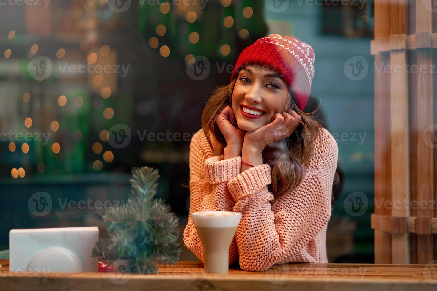 Beautiful caucasian blonde woman in red knitted hat sitting behind cafeteria window drinking a warm latte on a wonderful Christmas day. photo