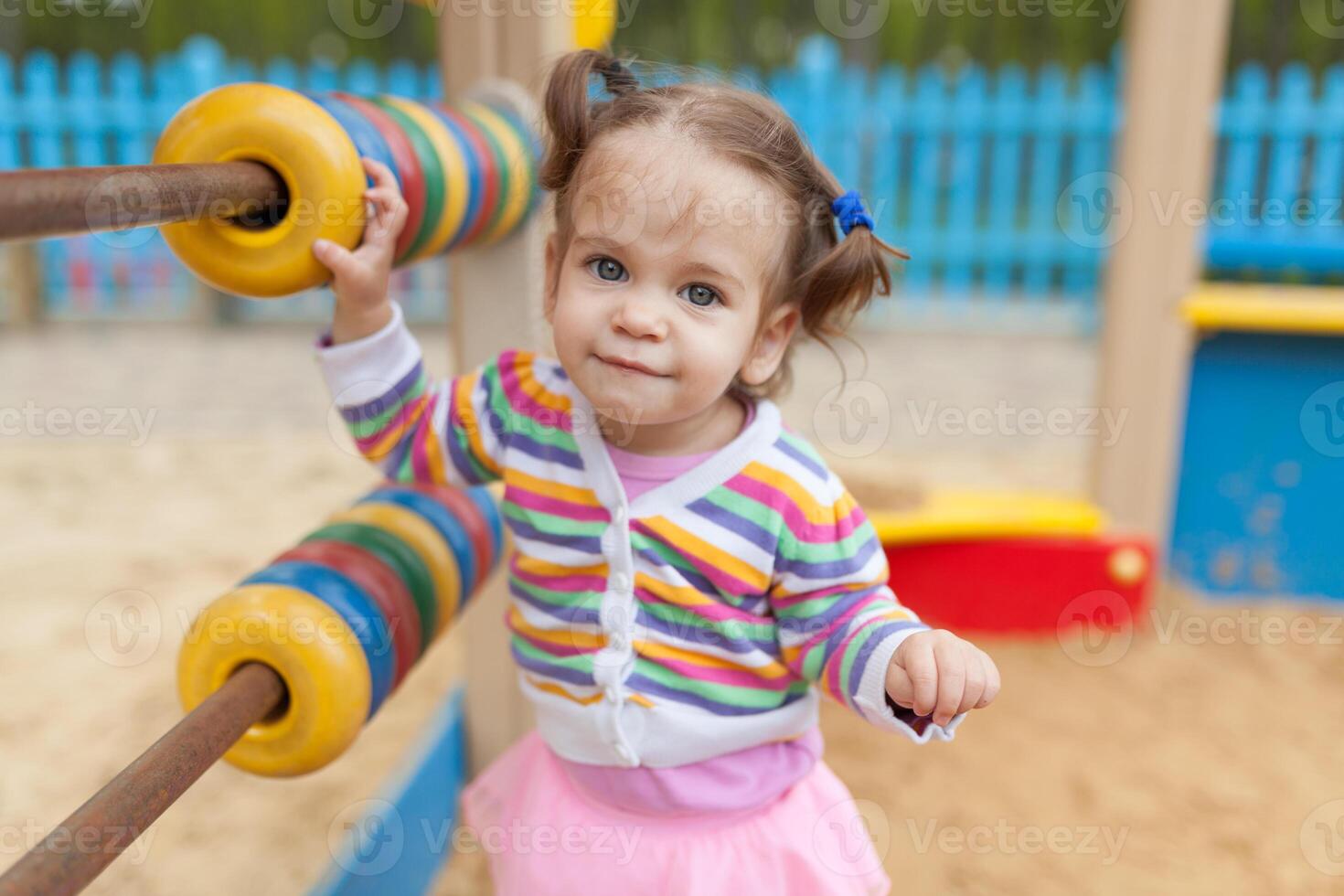 a little girl with two tails is dressed in a striped colorful jacket is playing in the sandbox on the playground photo