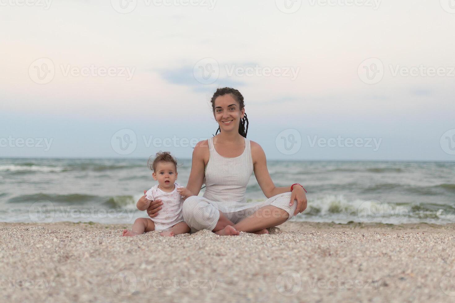 Happy family mother and child daughter doing yoga, meditate in lotus position on beach at sunset photo