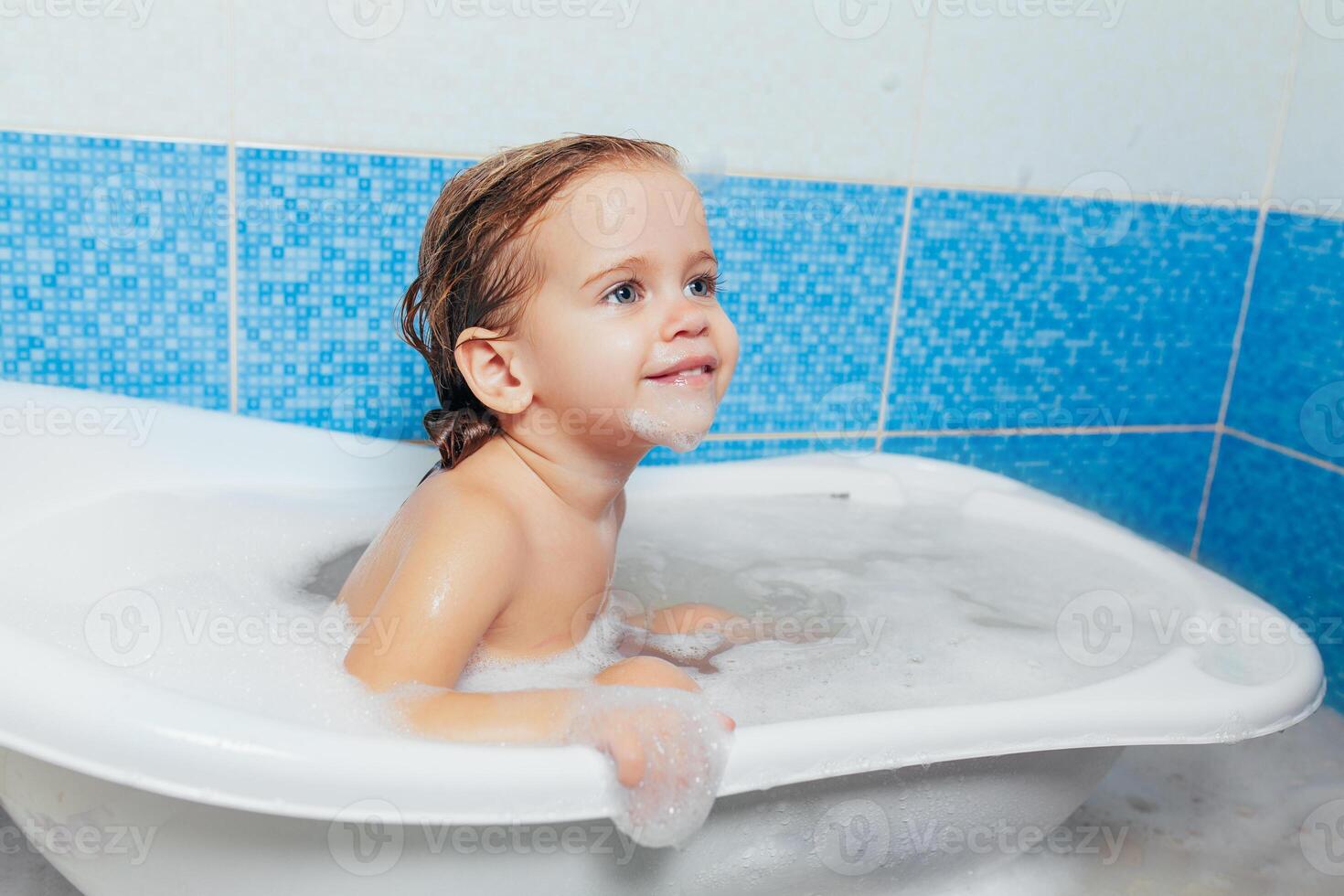 Fun cheerful happy toddler baby taking a bath playing with foam bubbles. Little child in a bathtub. Smiling kid in bathroom on blue background. Hygiene and health care. photo