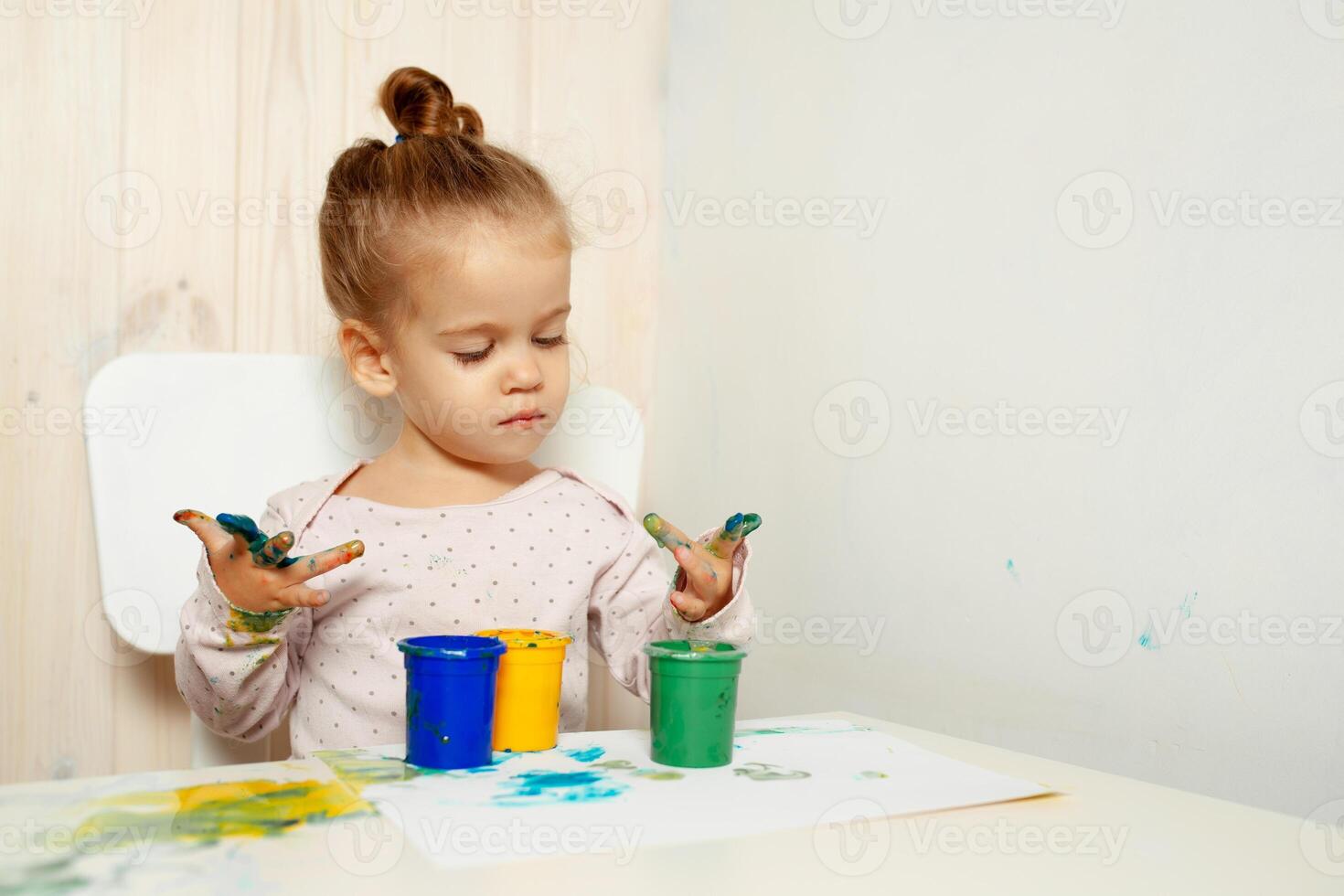 hermosa pequeño niña sorteos con dedo pinturas en un blanco sábana de papel. creativo niño desarrollo en jardín de infancia o gratis hora a hogar foto