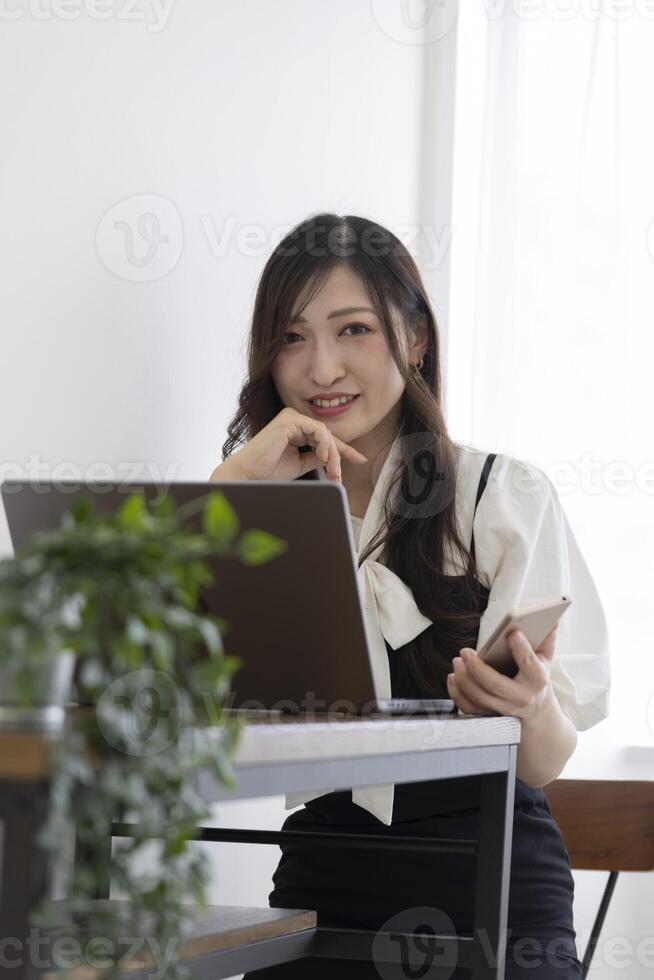 un japonés mujer comprobación teléfono inteligente por remoto trabajo en el pequeño oficina foto