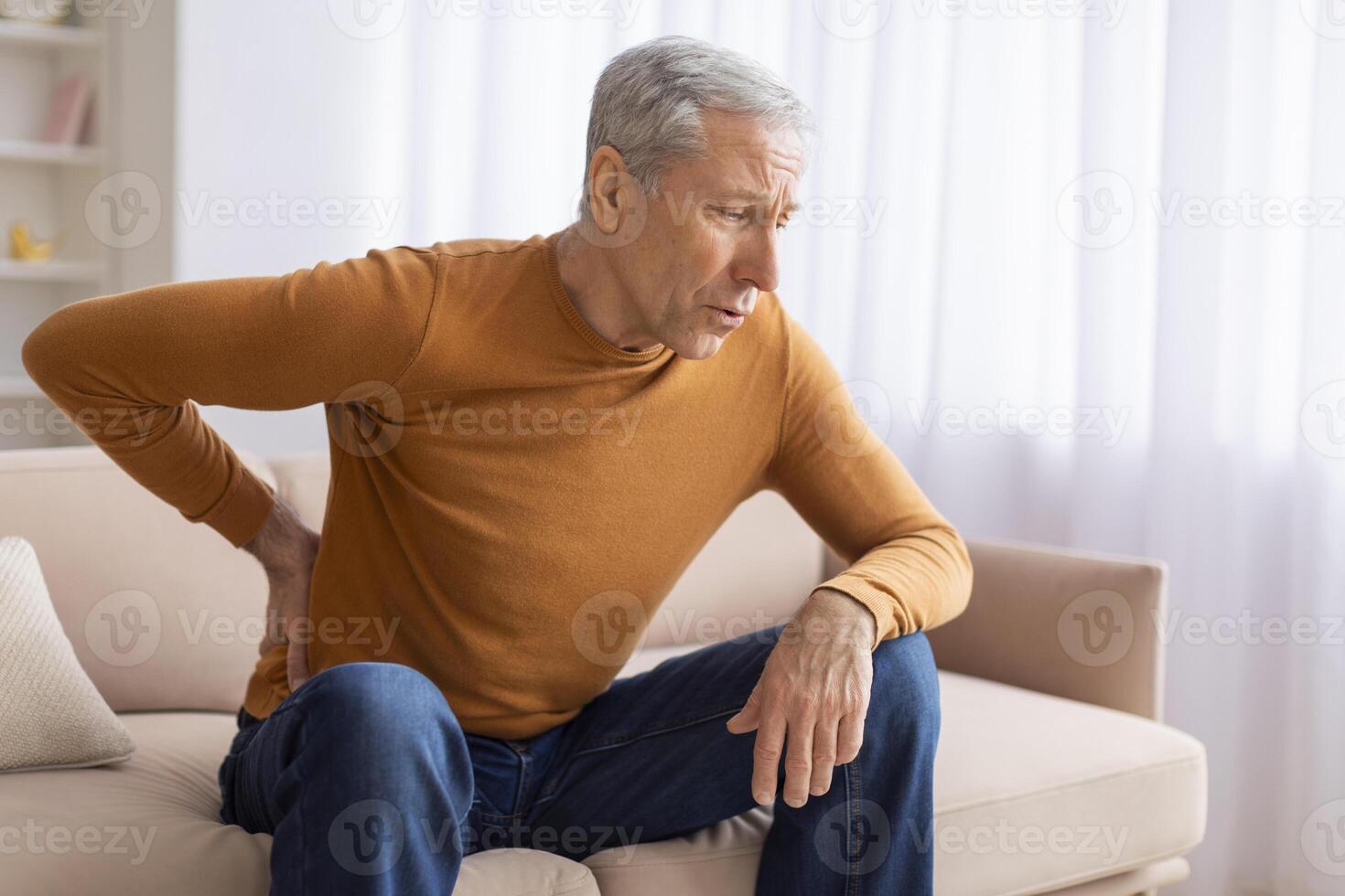 Unhappy grandfather sitting on couch and rubbing his lower back photo
