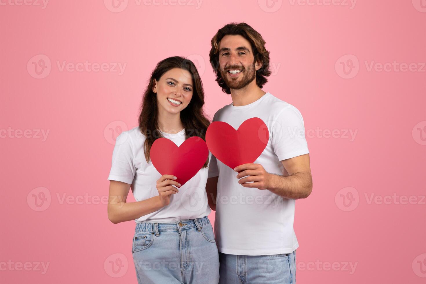 Happy couple in white t-shirts and jeans holding red heart-shaped paper cutouts photo