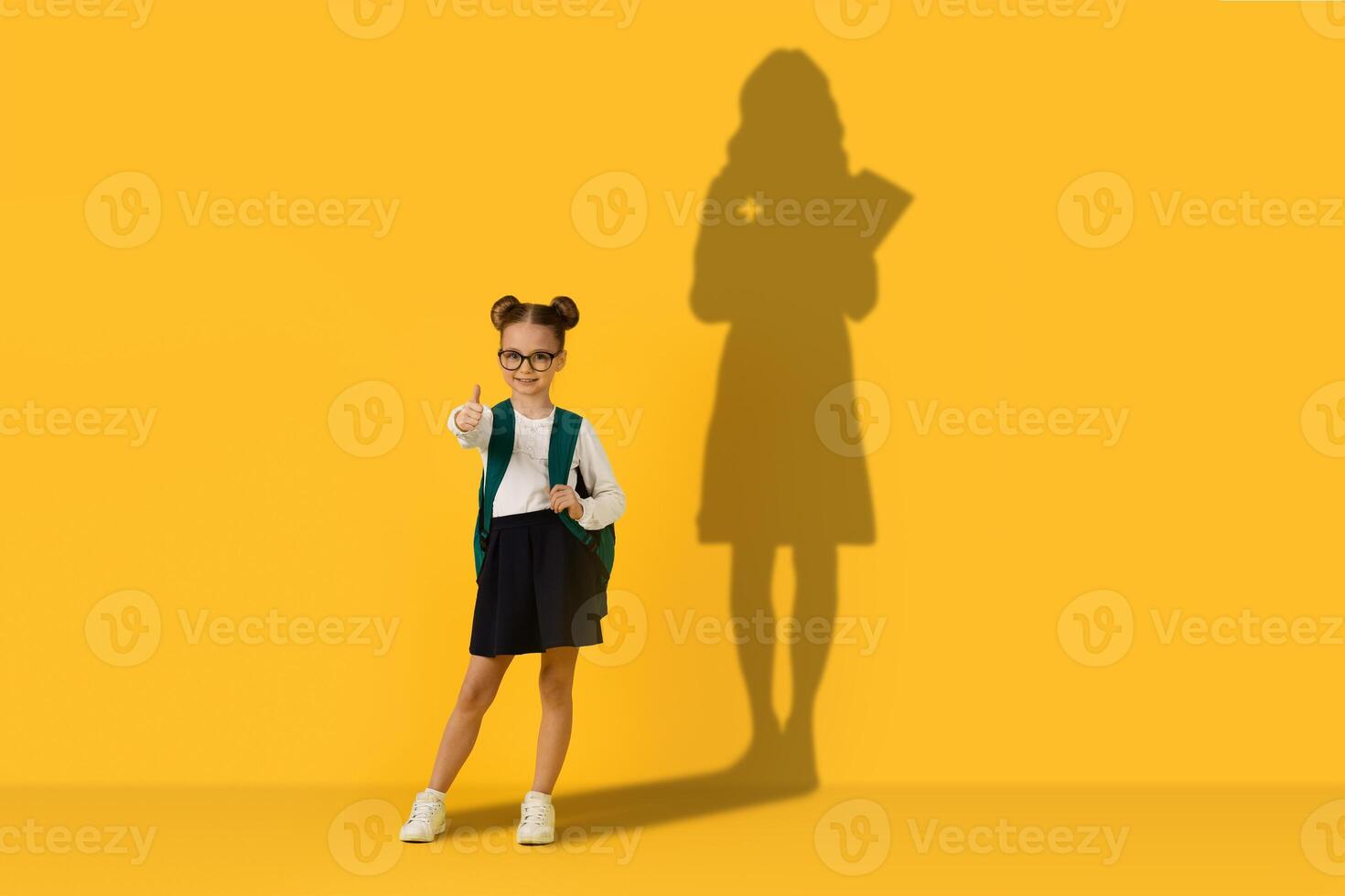 A confident schoolgirl with glasses and hair buns gives a thumbs up while holding a green backpack photo