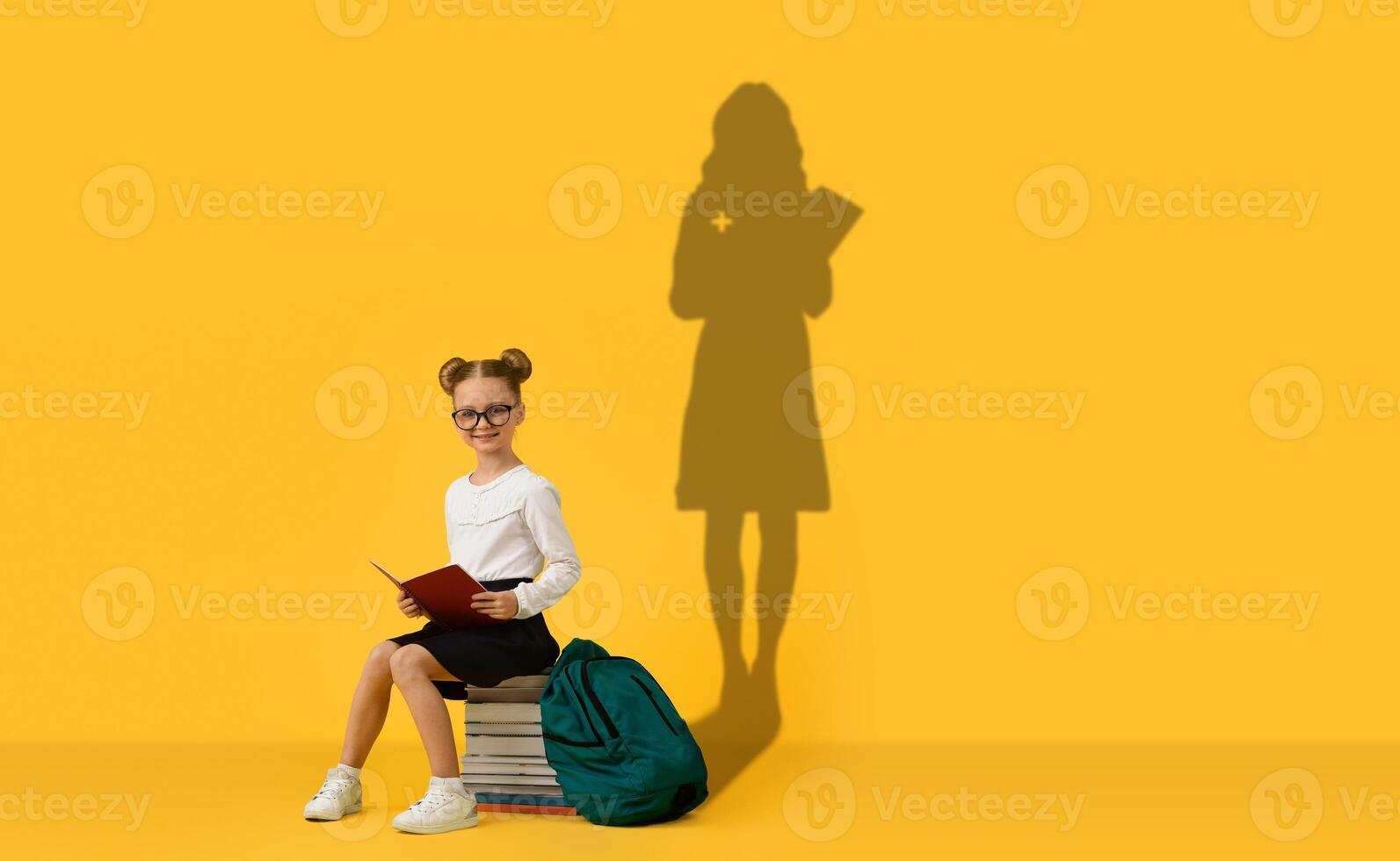 A young girl with glasses and hair in buns sits on a pile of books with a green backpack photo