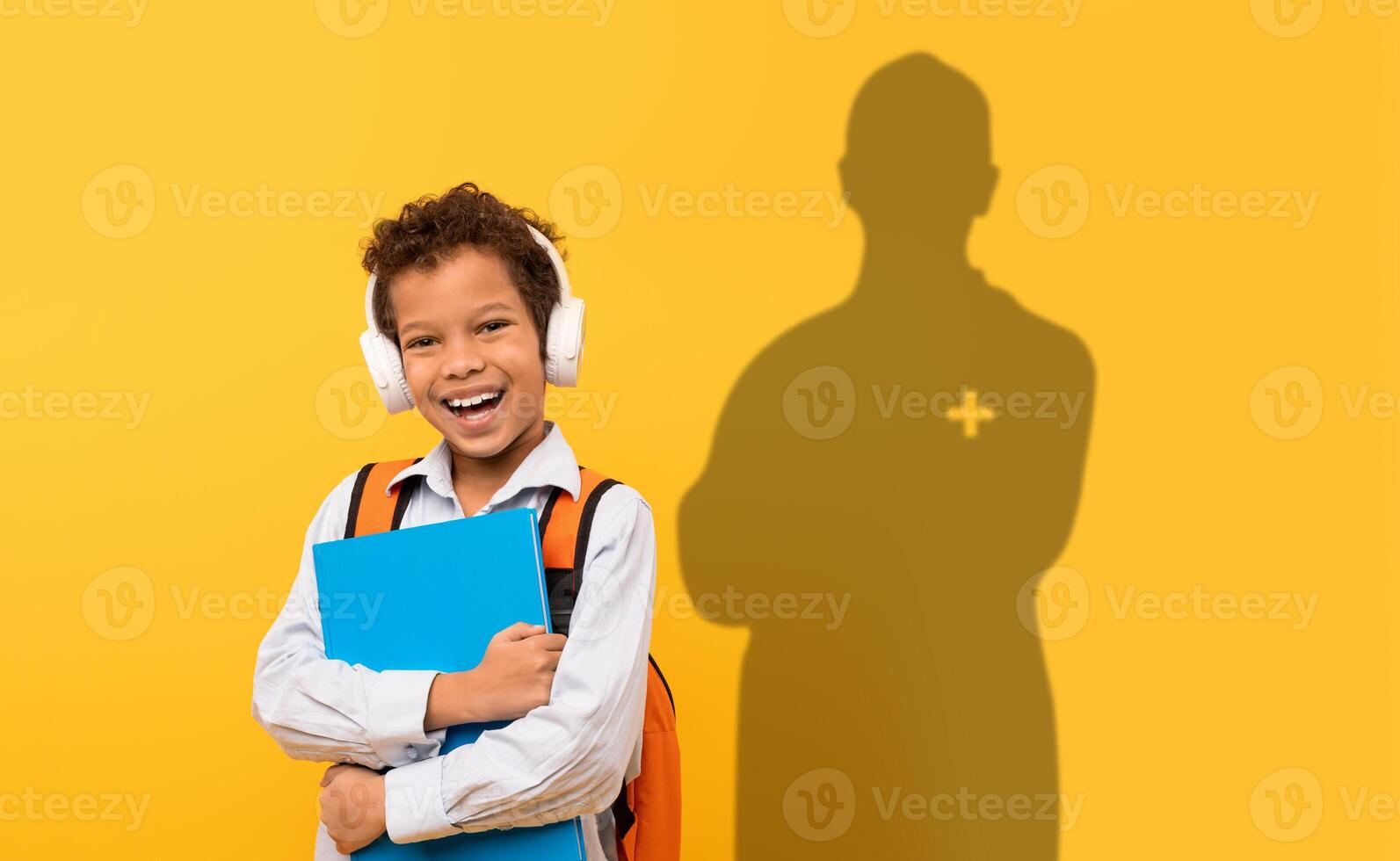 A beaming boy with headphones and an orange backpack hugs his blue folder photo