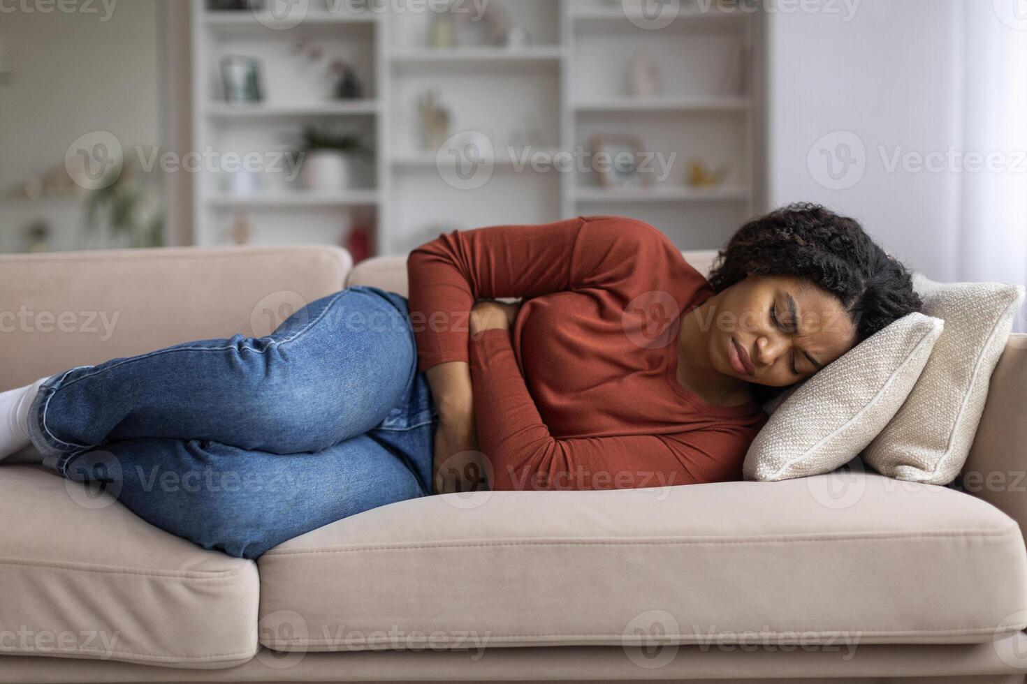 joven negro mujer agarrando su abdomen en incomodidad a hogar foto
