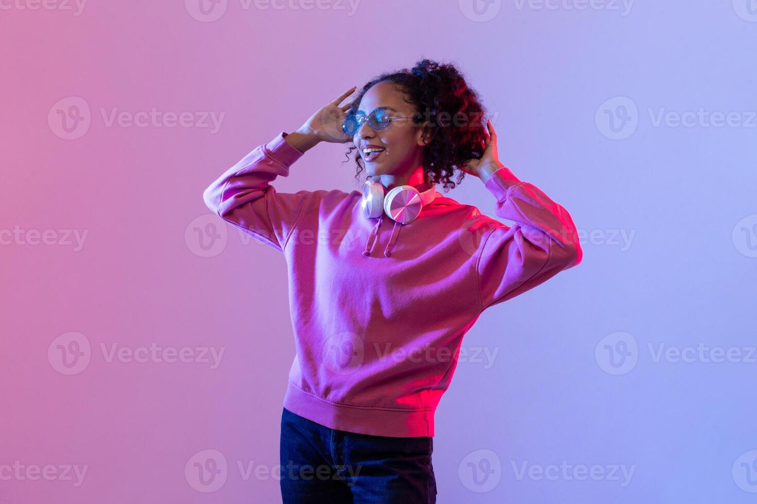 Joyful black woman with curly hair enjoying music with headphones in pink hoodie against neon background photo