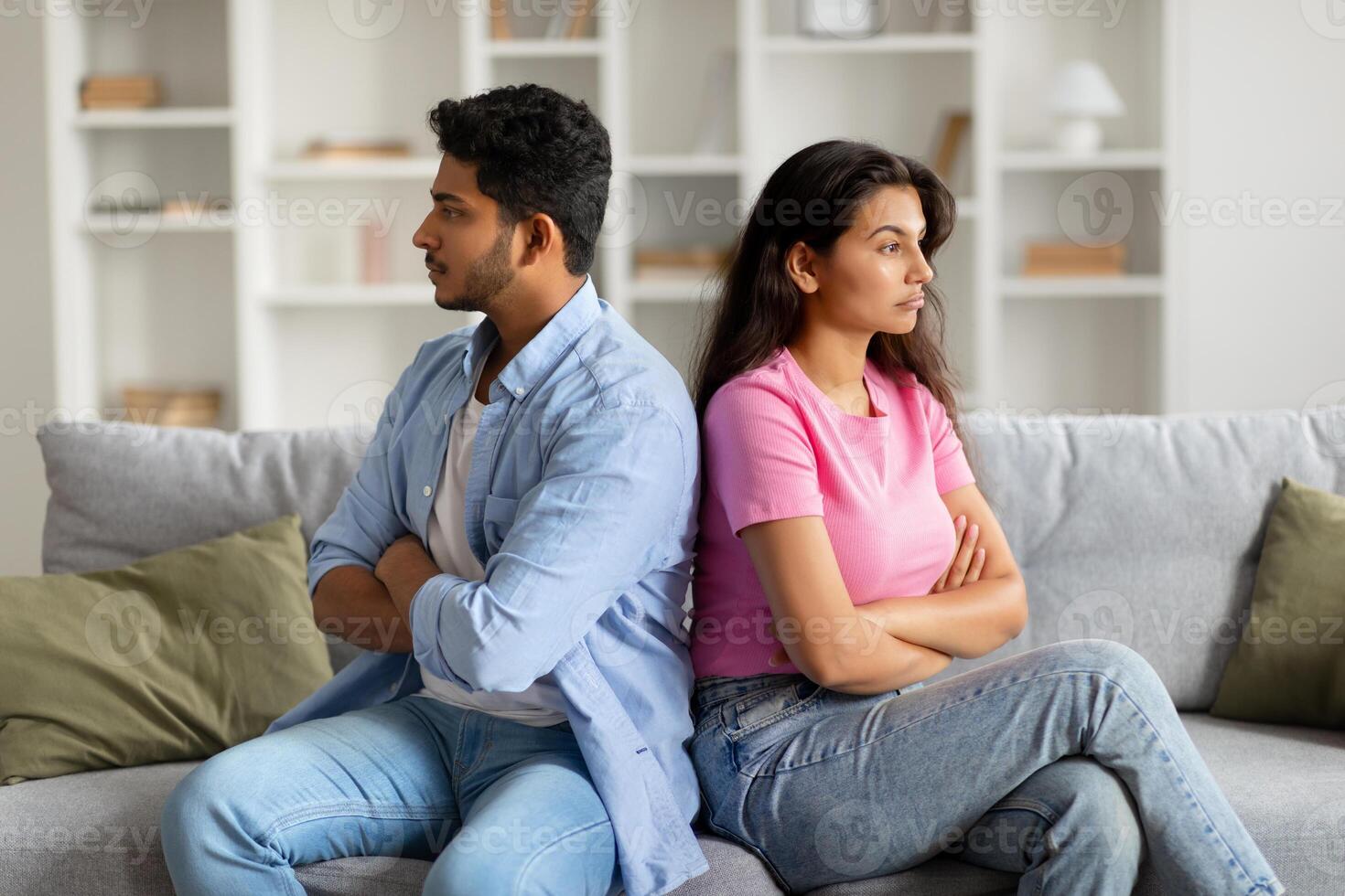 Unhappy indian couple sitting apart on sofa with arms crossed photo