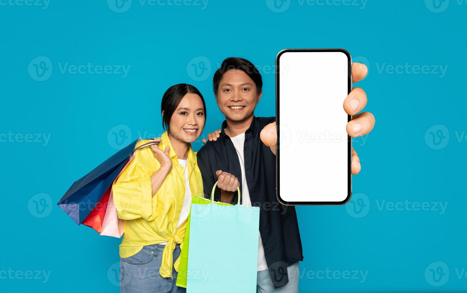 un sonriente hombre y mujer, que lleva vistoso compras bolsas, alegremente presente un teléfono inteligente con un blanco pantalla foto