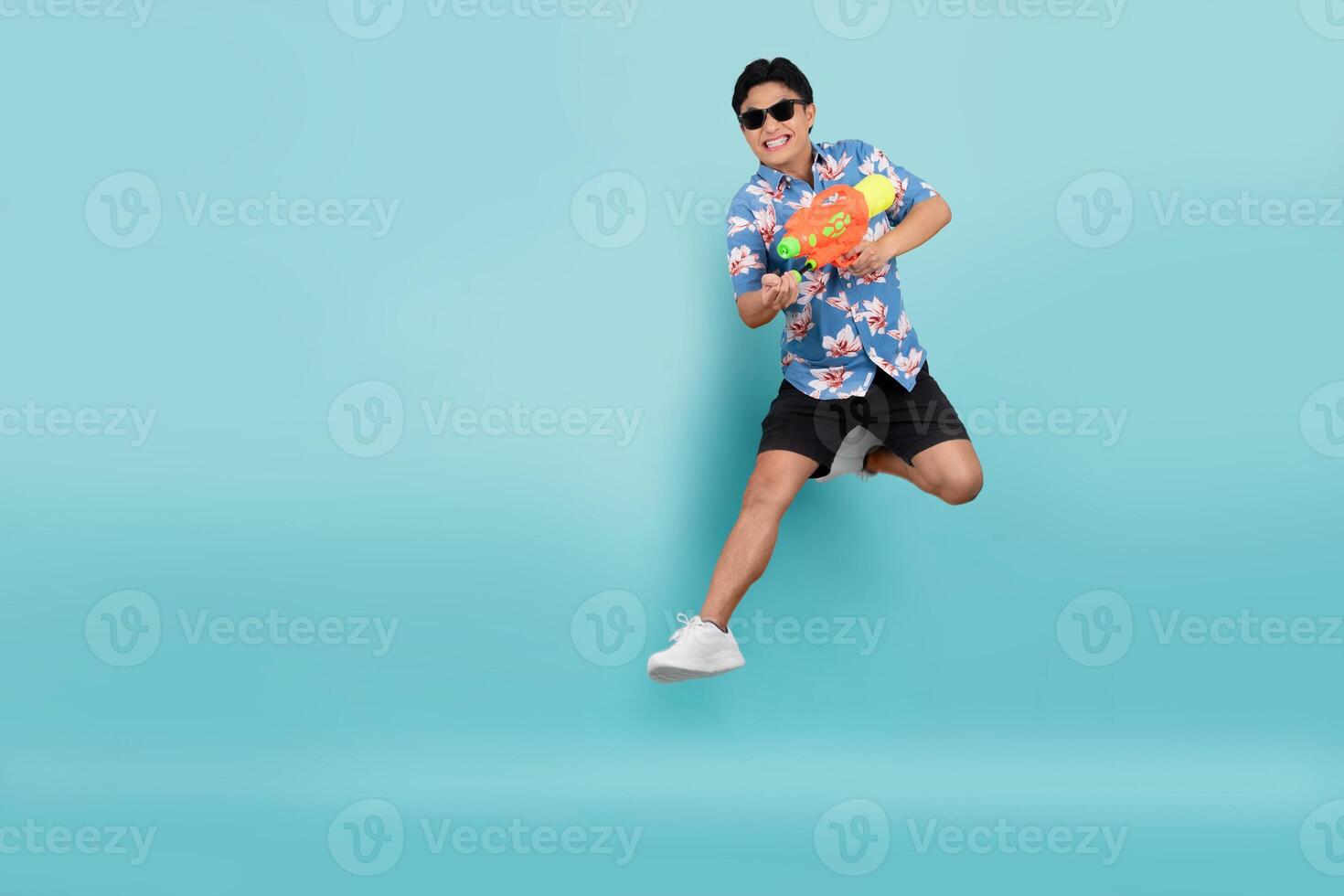 hermoso asiático joven hombre en verano ropa es saltando y utilizando un agua pistola en un azul antecedentes. Songkran festival en Tailandia foto