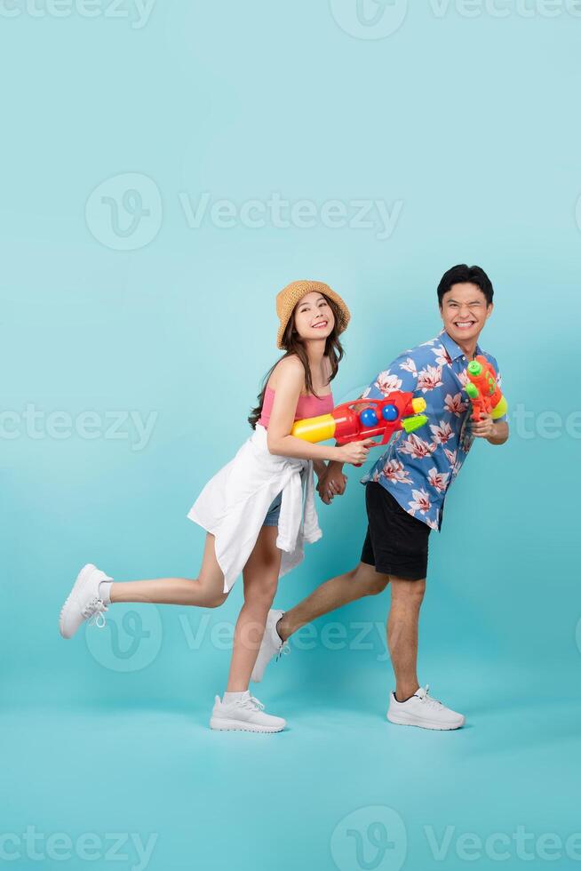 Side view of full length portrait of Young Asian couple walking and holding water gun isolated on blue background photo