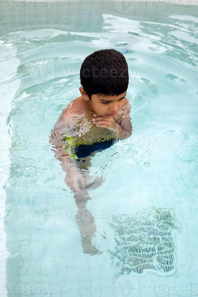 Happy Indian boy swimming in a pool, Kid wearing swimming costume along with air tube during hot summer vacations, Children boy in big swimming pool. photo