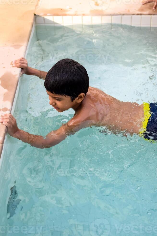 Happy Indian boy swimming in a pool, Kid wearing swimming costume along with air tube during hot summer vacations, Children boy in big swimming pool. photo